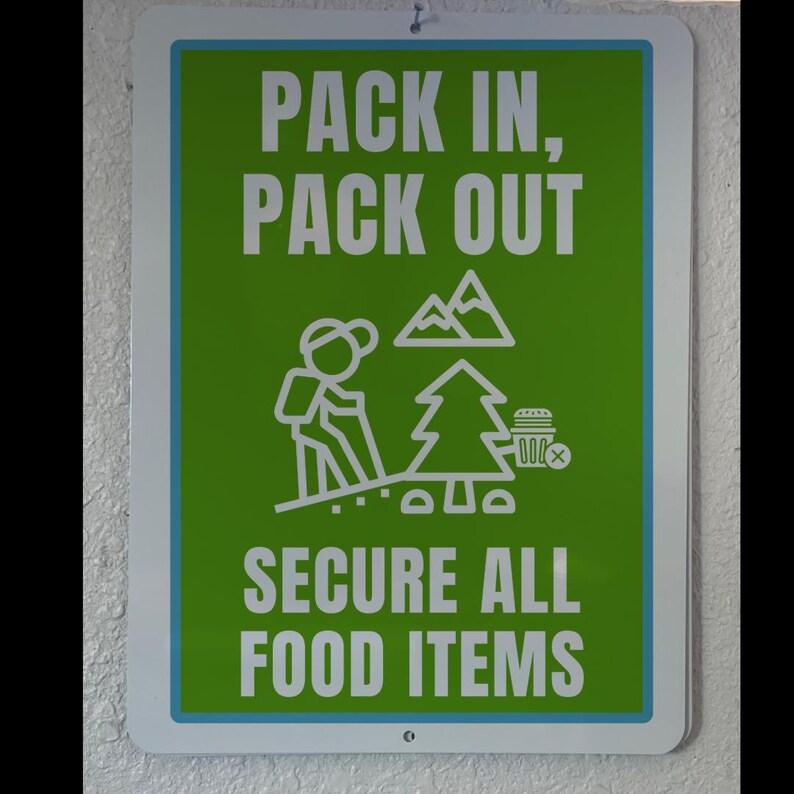 May include: Green sign with white text that reads "Pack in, Pack out" and "Secure all food items". A stick figure hiker is walking up a hill with a mountain and a tree in the background. A small white box with a red X is next to the tree.