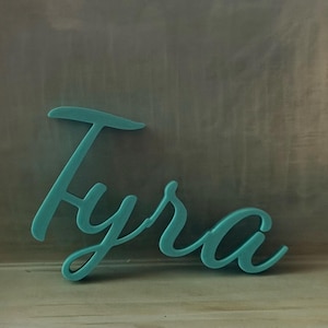 May include: A teal-colored, three-dimensional script sign featuring the name Tyra in a cursive font. The letters are connected in a single piece, displaying a smooth, matte finish and a consistent thickness throughout the entire word structure.