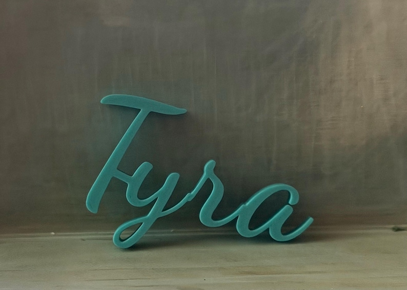 May include: A teal-colored, three-dimensional script sign featuring the name Tyra in a cursive font. The letters are connected in a single piece, displaying a smooth, matte finish and a consistent thickness throughout the entire word structure.