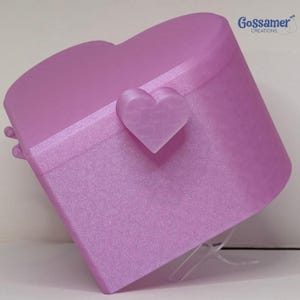 May include: A heart-shaped lavender storage box with a smooth, shimmering finish. The lid features a central heart-shaped latch and a horizontal band detail. The text Gossamer CREATIONS is printed in blue in the upper right corner of the image.