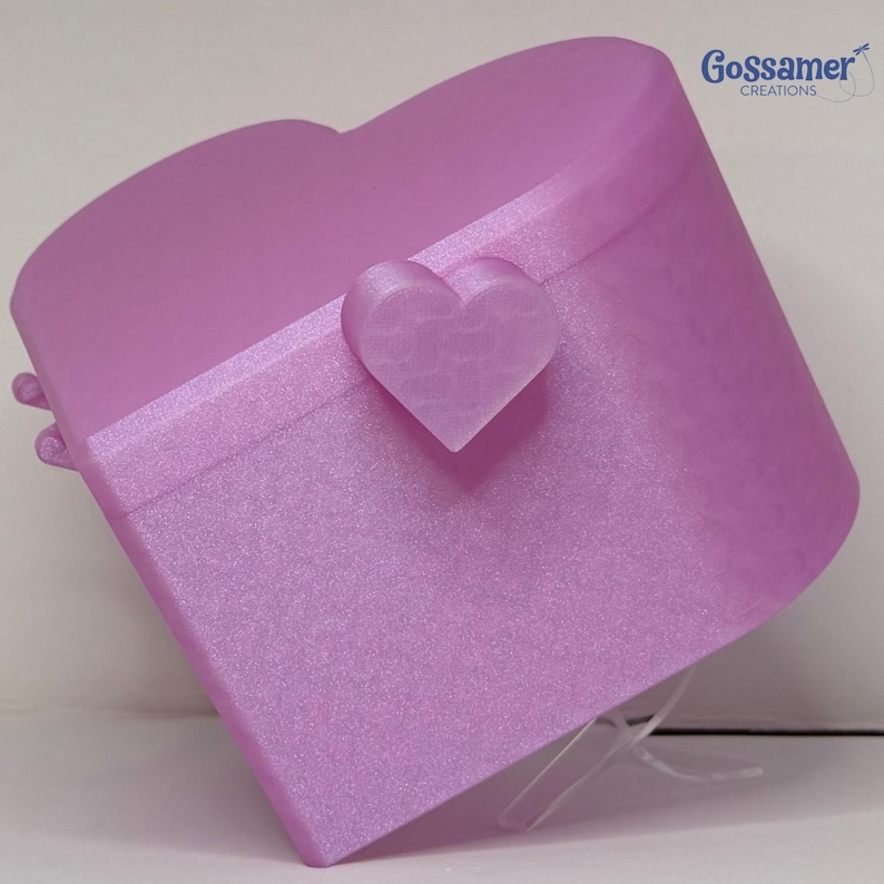 May include: A heart-shaped lavender storage box with a smooth, shimmering finish. The lid features a central heart-shaped latch and a horizontal band detail. The text Gossamer CREATIONS is printed in blue in the upper right corner of the image.