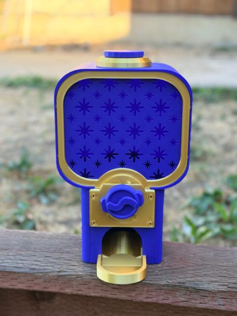 Op de afbeelding: Een blauwe en gouden kauwgomballenautomaat met een vierkante houder voorzien van een sterrenpatroon op het voorpaneel. Het apparaat heeft een ronde deksel aan de bovenkant, een centrale draaiknop en een klein gouden bakje onderaan.