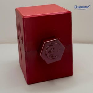 May include: A red, cube-shaped box with a hexagonal clasp. The clasp features a stylized emblem. The box has a glossy finish and is likely made of metal. The upper right corner of the image shows the text "Gossamer Creations".