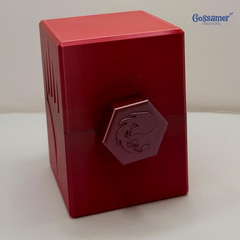 May include: A red, cube-shaped box with a hexagonal clasp. The clasp features a stylized emblem. The box has a glossy finish and is likely made of metal. The upper right corner of the image shows the text "Gossamer Creations".