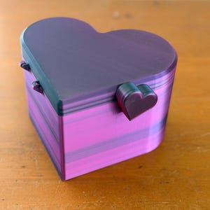May include: A heart-shaped box with a lid, featuring a gradient of dark purple and vibrant magenta horizontal stripes. The box has a small heart-shaped latch on the front and two visible hinges on the back, constructed with a layered texture.