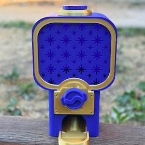 May include: A blue and gold gumball machine featuring a square container with a repeating star pattern on the front panel. The device includes a gold-coloured dispensing mechanism with a blue knob and a small gold tray at the base for item collection.