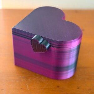 May include: A heart-shaped box with a lid, featuring a gradient of purple and black horizontal stripes. The box has a small, dark, heart-shaped latch on the front. The surface shows a fine, layered texture consistent with 3D printed objects.