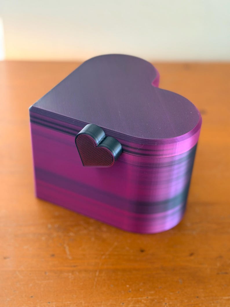 May include: A heart-shaped box with a lid, featuring a gradient of purple and black horizontal stripes. The box has a small, dark, heart-shaped latch on the front. The surface shows a fine, layered texture consistent with 3D printed objects.