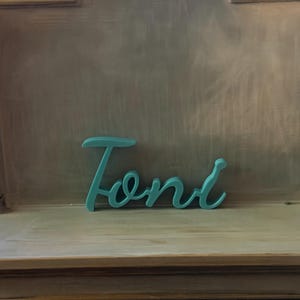May include: A teal-colored, three-dimensional script sign featuring the name Toni in a cursive font. The letters are connected in a single piece, displaying a smooth, matte finish and a consistent thickness throughout the entire word structure.