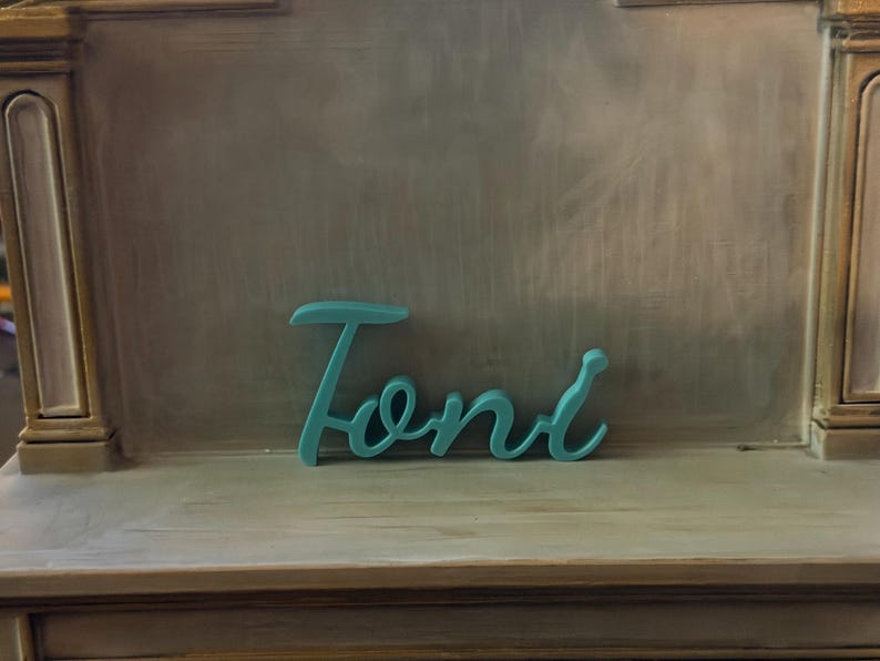 May include: A teal-colored, three-dimensional script sign featuring the name Toni in a cursive font. The letters are connected in a single piece, displaying a smooth, matte finish and a consistent thickness throughout the entire word structure.