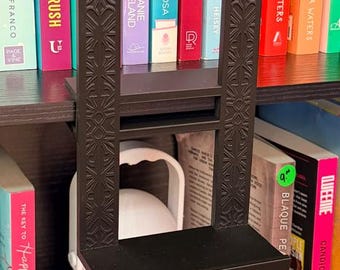 May include: A black, rectangular, 3D-printed shelf designed to hook onto the top of a bookshelf. The vertical side supports feature an intricate, symmetrical, geometric, embossed pattern, while the horizontal shelf provides a flat surface.