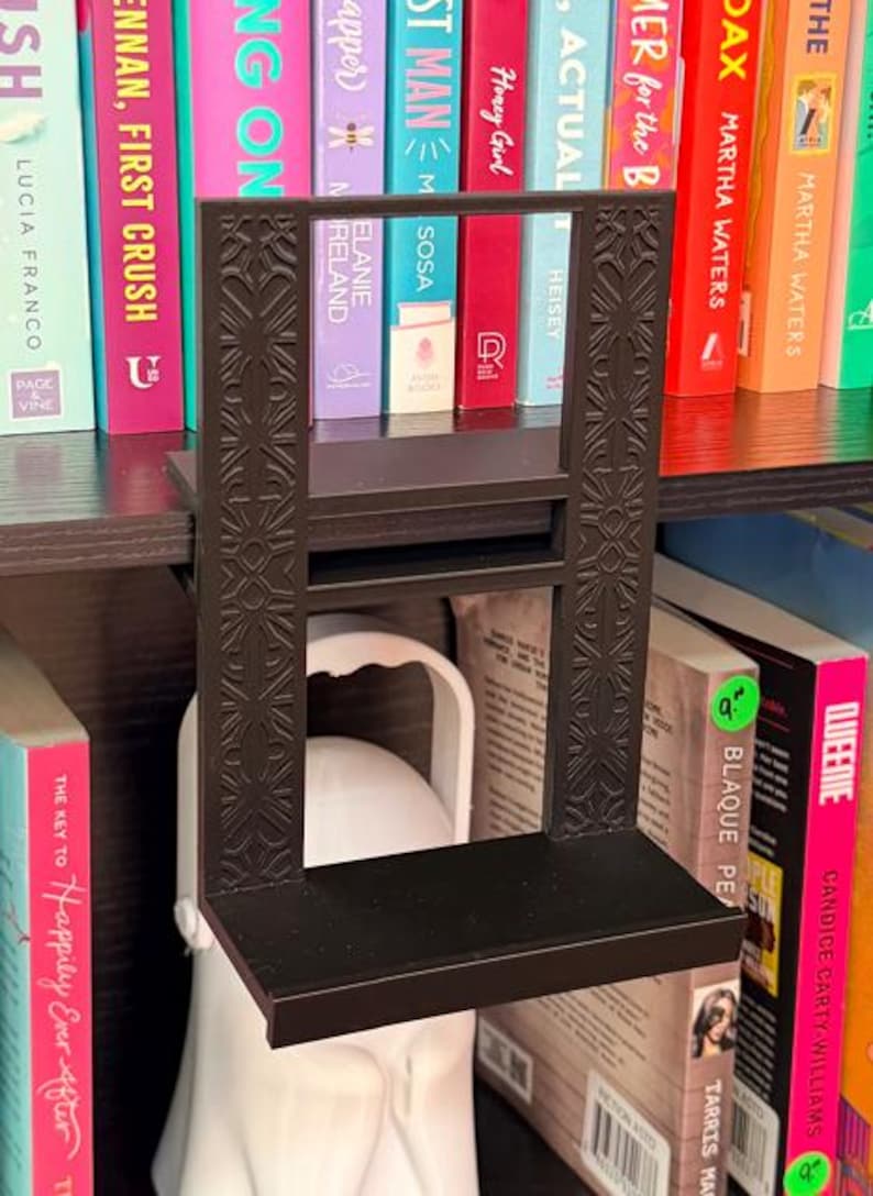 K&ouml;nnte beinhalten: Ein schwarzes, rechteckiges, 3D-gedrucktes Regal, das zum Einh&auml;ngen an die Oberseite eines B&uuml;cherregals konzipiert ist. Die vertikalen Seitenst&uuml;tzen weisen ein komplexes, symmetrisches, geometrisches Pr&auml;gemuster auf, w&auml;hrend das horizontale Regal eine flache Ablagefl&auml;che bietet.