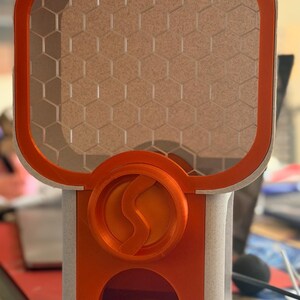 May include: A 3D printed dispenser featuring a bright orange frame and a light beige, textured body. The upper section has a honeycomb grid pattern, while the centre displays a circular orange emblem with a stylised S-shaped logo in relief.