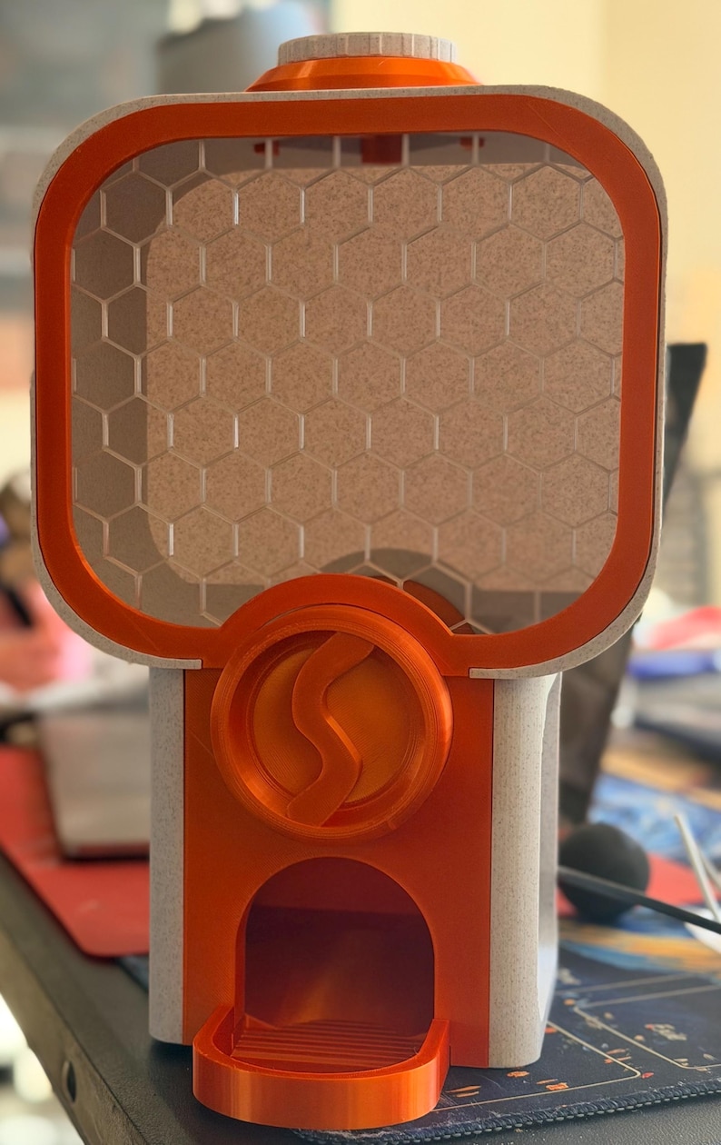 May include: A 3D printed dispenser featuring a bright orange frame and a light beige, textured body. The upper section has a honeycomb grid pattern, while the centre displays a circular orange emblem with a stylised S-shaped logo in relief.
