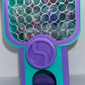 May include: A purple and teal 3D-printed gumball machine featuring a honeycomb-patterned front panel filled with clear plastic capsules containing small items. The machine has a central circular dial and a bottom dispensing tray.