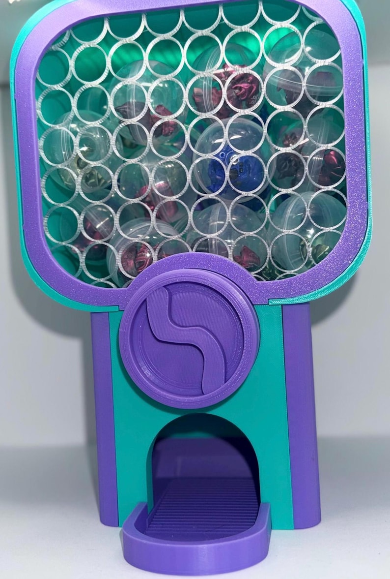 May include: A purple and teal 3D-printed gumball machine featuring a honeycomb-patterned front panel filled with clear plastic capsules containing small items. The machine has a central circular dial and a bottom dispensing tray.