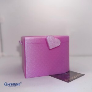 May include: A shimmering magenta box with a rounded front and a matching heart-shaped latch on the lid. The surface has a textured, glittery appearance. A small dark rectangular card with the text Gossamer Creations is placed nearby.