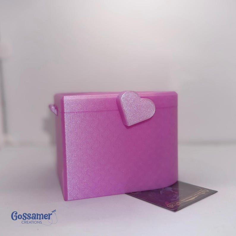 May include: A shimmering magenta box with a rounded front and a matching heart-shaped latch on the lid. The surface has a textured, glittery appearance. A small dark rectangular card with the text Gossamer Creations is placed nearby.