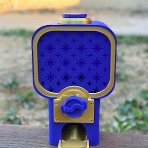 Op de afbeelding: Een blauwe en gouden kauwgomballenautomaat met een rechthoekige container met een sterrenpatroon op het voorpaneel. Het apparaat bevat een goudkleurig doseermechanisme, een ronde deksel aan de bovenkant en een kleine gouden opvangbak.