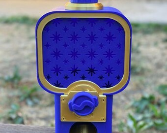 May include: A blue and gold gumball machine featuring a rectangular container with a star pattern on the front panel. The device includes a gold-colored dispensing mechanism, a circular top lid, and a small gold tray at the base for items.