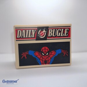 May include: A rectangular, off-white box featuring the Daily Bugle logo in red and black. Below the logo is a black rectangle with a red and blue Spider-Man graphic. The box has a glossy finish.