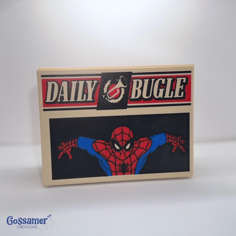 May include: A rectangular, off-white box featuring the Daily Bugle logo in red and black. Below the logo is a black rectangle with a red and blue Spider-Man graphic. The box has a glossy finish.