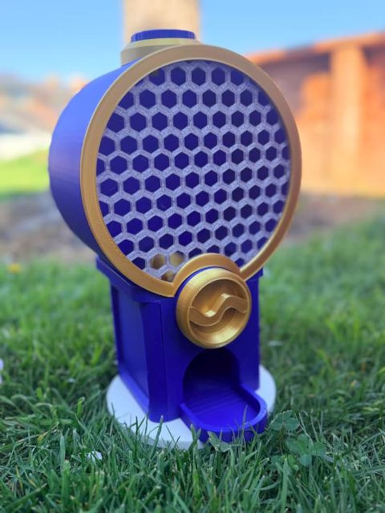 May include: A blue and gold gumball machine featuring a circular container with a honeycomb pattern. The device has a gold-colored dispensing knob, a blue base, and a small chute at the bottom for dispensing items. The structure is upright and cylindrical.