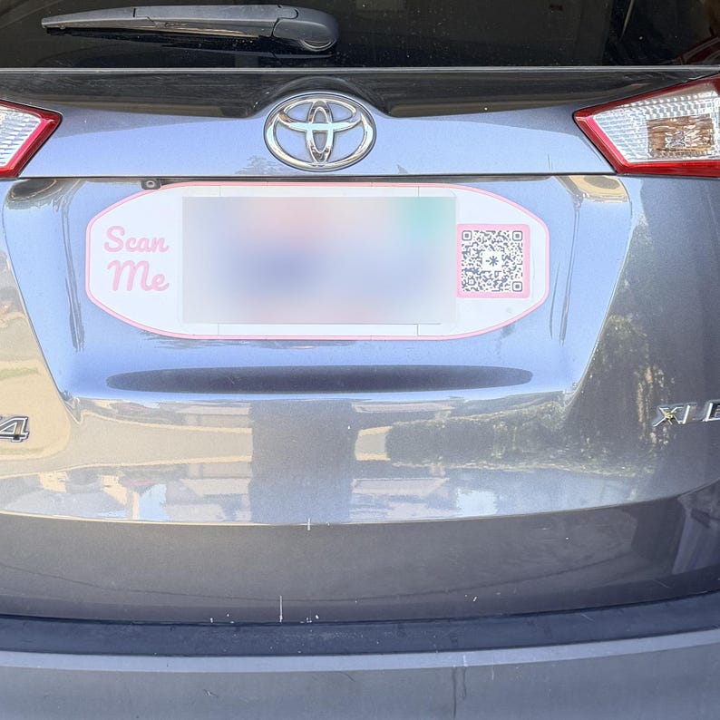 May include: A grey Toyota vehicle rear hatch featuring a custom oval number plate frame. The frame has a pink border, the text Scan Me in pink script on the left side, and a black and white QR code on the right side of the plate area.