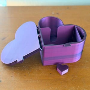 May include: A heart-shaped purple storage box with a hinged lid and a small, separate heart-shaped piece. The container features a divided interior with a cutout section on one of the inner walls, showcasing a smooth, layered finish throughout.