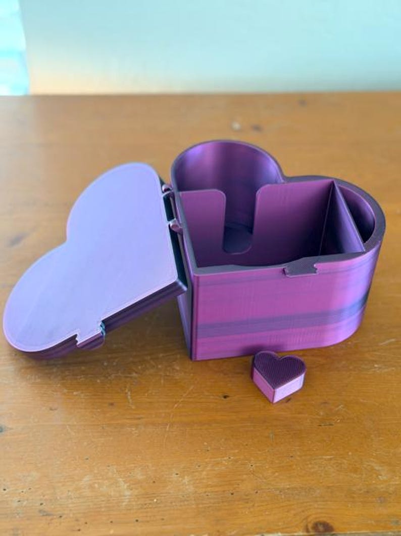 May include: A heart-shaped purple storage box with a hinged lid and a small, separate heart-shaped piece. The container features a divided interior with a cutout section on one of the inner walls, showcasing a smooth, layered finish throughout.