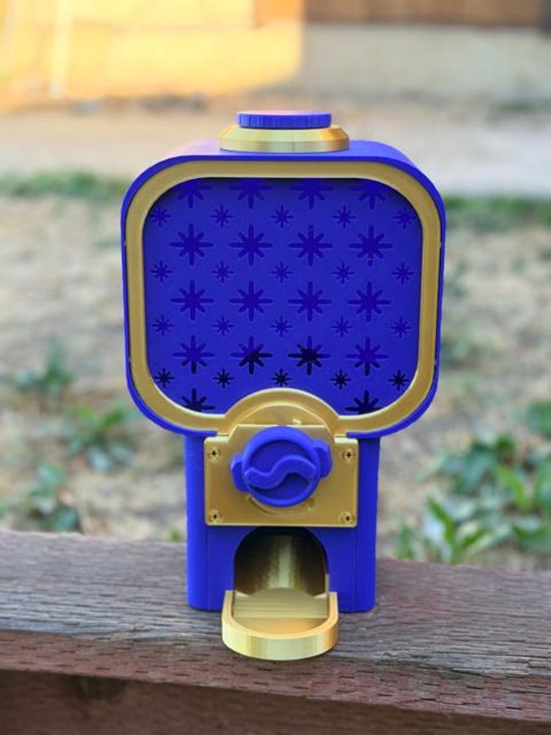 Op de afbeelding: Een blauwe en gouden kauwgomballenautomaat met een vierkante houder voorzien van een sterrenpatroon op het voorpaneel. Het apparaat bevat een ronde blauwe doseerknop, een gouden rechthoekige voorplaat en een gouden opvangbakje aan de basis.