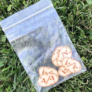 May include: A clear plastic resealable bag containing five small, irregular, cream-colored stickers. Each sticker features a brown graphic of a stylized face with a line for a mouth and two small dots for eyes, surrounded by tiny star-like accents.