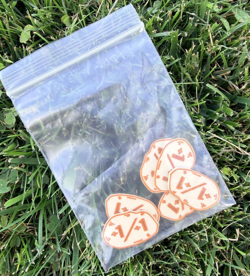 K&ouml;nnte beinhalten: Ein durchsichtiger, wiederverschlie&szlig;barer Plastikbeutel mit f&uuml;nf kleinen, unregelm&auml;&szlig;ig geformten, cremefarbenen Aufklebern. Jeder Aufkleber zeigt eine braune Grafik eines stilisierten Gesichts mit einem Strich als Mund und zwei Punkten als Augen.