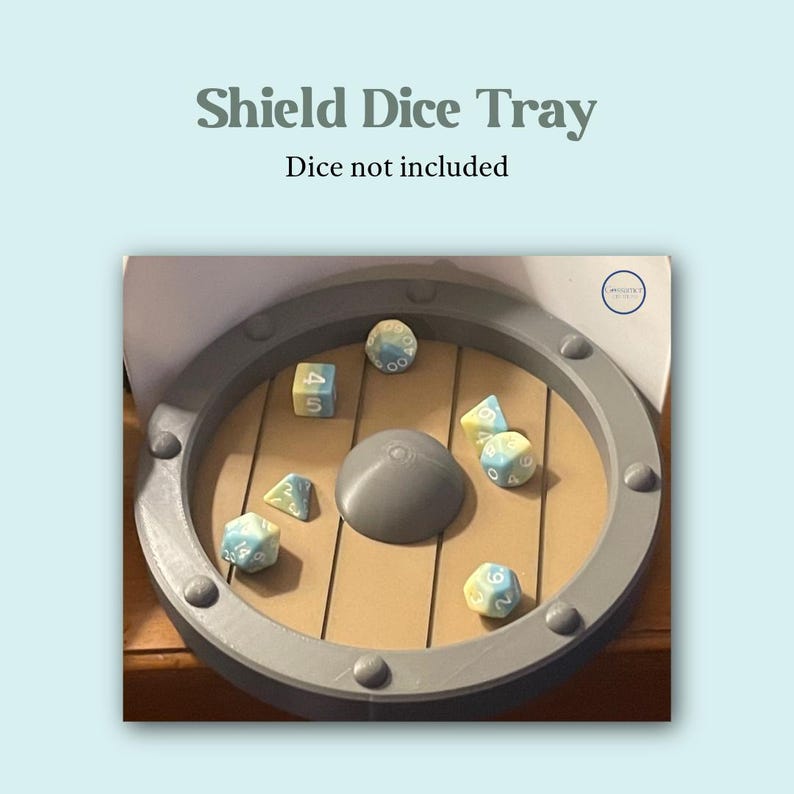 May include: A gray shield-shaped dice tray with a wooden interior and dice. The tray has a gray rim with rivets and a central gray dome. The text "Shield Dice Tray" and "Dice not included" are at the top.