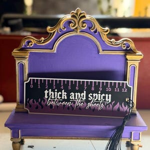 May include: A purple and gold decorative wooden stand featuring a ruler graphic with the text thick and spicy between the sheets. The ruler shows 12 inches. A black tassel hangs from the right side of the rectangular sign.