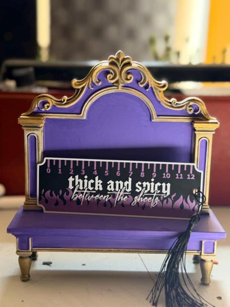 May include: A purple and gold decorative wooden stand featuring a ruler graphic with the text thick and spicy between the sheets. The ruler shows 12 inches. A black tassel hangs from the right side of the rectangular sign.