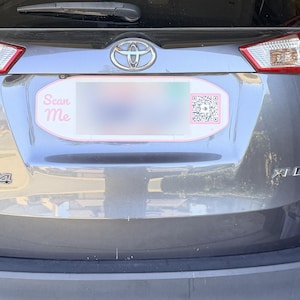 May include: A grey Toyota RAV4 XLE vehicle featuring a custom oval number plate frame with a red border. The frame includes the text Scan Me in pink lettering on the left side and a black and white QR code on the right side of the plate area.
