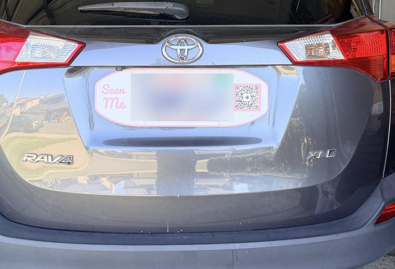 May include: A grey Toyota RAV4 XLE vehicle featuring a custom oval number plate frame with a red border. The frame includes the text Scan Me in pink lettering on the left side and a black and white QR code on the right side of the plate area.