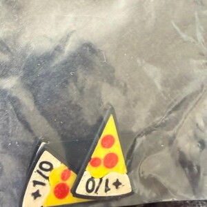 May include: Two triangular pizza slice pins with yellow cheese, red pepperoni, and a white crust section. Each pin features the black text +1/0 printed on the white crust area. The pins have a dark border and are encased in a clear plastic bag.