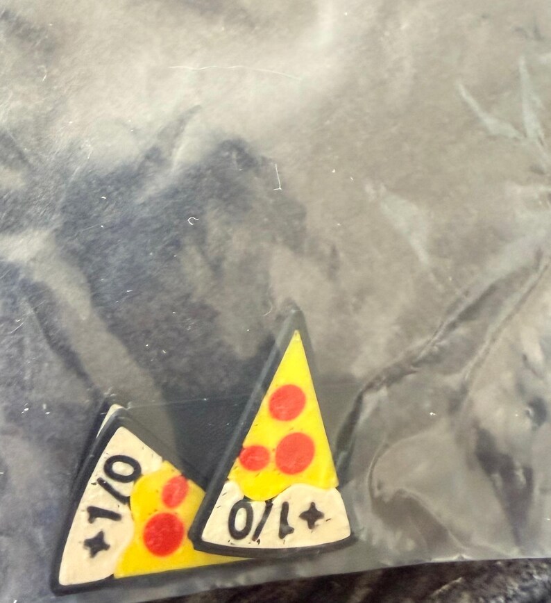 May include: Two triangular pizza slice pins with yellow cheese, red pepperoni, and a white crust section. Each pin features the black text +1/0 printed on the white crust area. The pins have a dark border and are encased in a clear plastic bag.