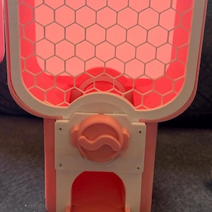 May include: A pink and white plastic gumball machine featuring a hexagonal grid pattern on the front panel, a central rotating knob, and a dispensing chute. A small clear plastic bag containing several light blue circular tokens is included.