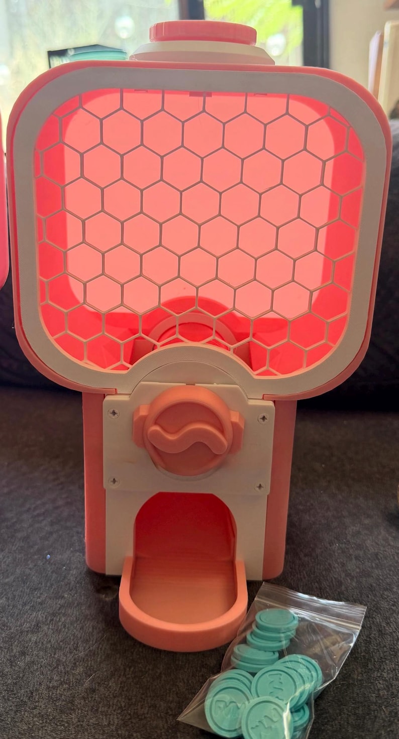 May include: A pink and white plastic gumball machine featuring a hexagonal grid pattern on the front panel, a central rotating knob, and a dispensing chute. A small clear plastic bag containing several light blue circular tokens is included.