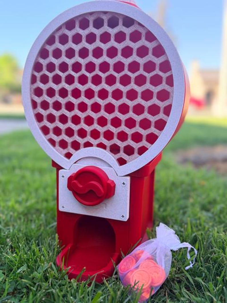 May include: A red and white gumball machine featuring a circular top section with a honeycomb pattern. The machine has a central red dispensing knob on a white square plate and a bottom chute. A small white organza bag filled with red tokens sits nearby.