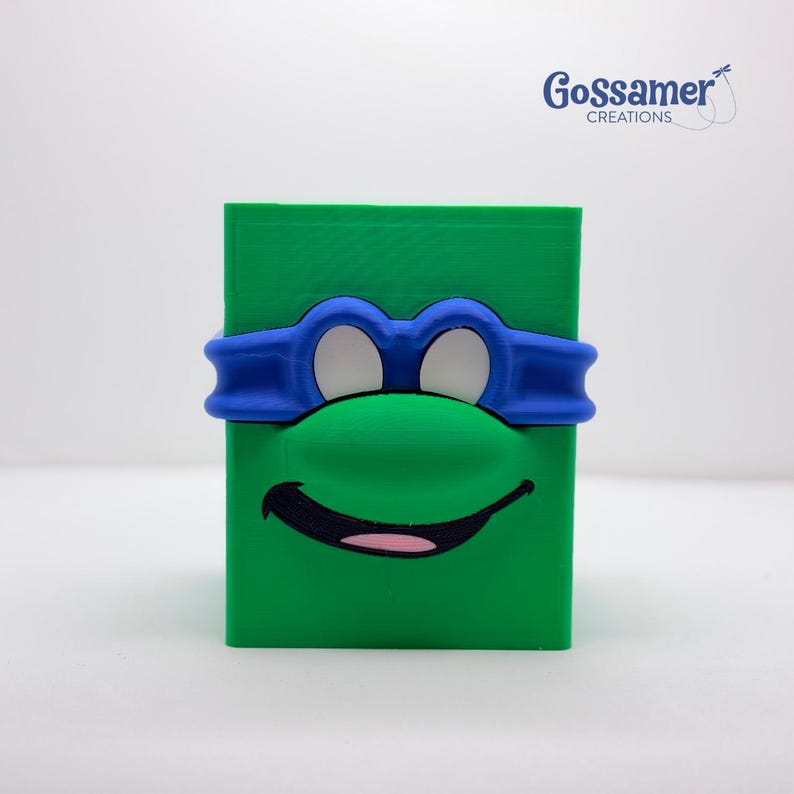 May include: A green, rectangular box with a blue mask and a green nose, resembling a cartoon character. The character's mouth is black with a pink tongue. The logo "Gossamer Creations" is in the upper right corner.