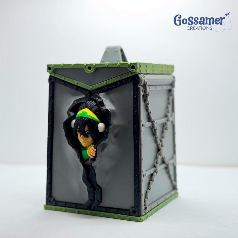 May include: A gray and green box-shaped container with a handle on top. The container features a character emerging from a torn section. The character has yellow and green headwear. The container has chain-like details on the sides.