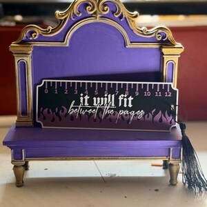 May include: A purple and gold display stand with an ornate arched top and four legs. Resting on the stand is a black bookmark featuring a ruler graphic from 0 to 12 inches, purple flame patterns, and the text "it will fit between the pages".