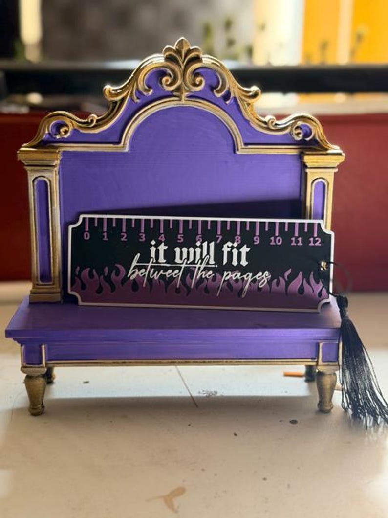 May include: A purple and gold display stand with an ornate arched top and four legs. Resting on the stand is a black bookmark featuring a ruler graphic from 0 to 12 inches, purple flame patterns, and the text "it will fit between the pages".