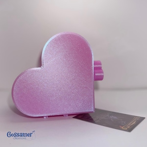 May include: A heart-shaped object with a shimmering light pink finish and a textured surface. On the right side, there are two small cylindrical protrusions. A small rectangular tag with the text Gossamer Creations is placed nearby.
