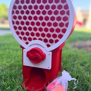 May include: A red and white gumball machine featuring a circular top section with a honeycomb pattern and a red dispensing knob. A small white organza drawstring bag filled with round red tokens sits next to the base of the machine.