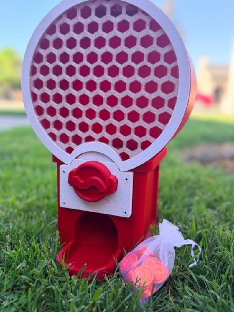 May include: A red and white gumball machine featuring a circular top section with a honeycomb pattern and a red dispensing knob. A small white organza drawstring bag filled with round red tokens sits next to the base of the machine.